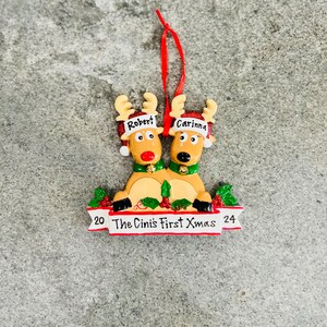 May include: A Christmas ornament featuring two reindeer with the names "Robert" and "Carinna" on their hats. The reindeer are tan with red noses and green collars. The ornament reads "20 The Cini's First Xmas 24" on a red banner.