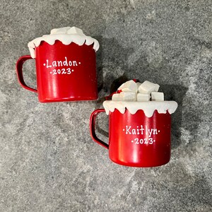 May include: Two red mugs with white trim and handles, topped with white foam and sugar cubes. One mug has "Landon" and "2023" in white text. The other mug has "Kaitlyn" and "2023" in white text.