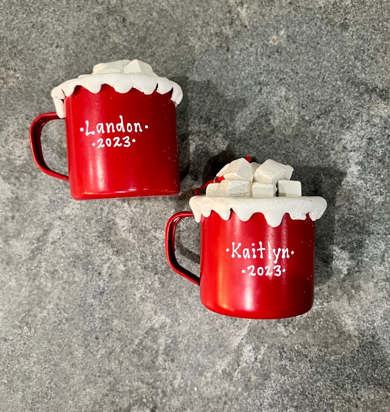 May include: Two red mugs with white trim and handles, topped with white foam and sugar cubes. One mug has "Landon" and "2023" in white text. The other mug has "Kaitlyn" and "2023" in white text.
