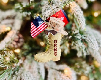 Puede incluir: Adorno navideño de bota USMC beige con las letras "USMC" en rojo. El adorno incluye un sombrero a juego y está adornado con una bandera estadounidense y otra bandera. El adorno cuelga de un árbol de Navidad cubierto de nieve.