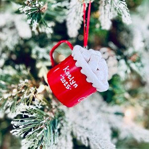 May include: A red Christmas ornament shaped like a mug, hanging on a snowy tree branch. The mug has a white, fluffy top and the name "Kaelyn" and the year "2025" written on it in white. A red ribbon is used to hang the ornament.