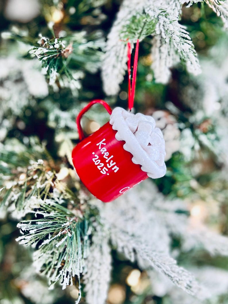 May include: A red Christmas ornament shaped like a mug, hanging on a snowy tree branch. The mug has a white, fluffy top and the name "Kaelyn" and the year "2025" written on it in white. A red ribbon is used to hang the ornament.
