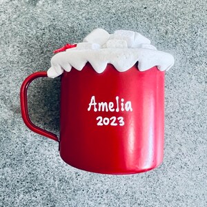May include: A red mug with a handle, topped with white frosting and marshmallows. The mug has the name "Amelia" and the year "2023" printed in white.