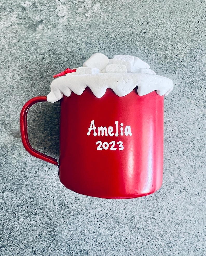 May include: A red mug with a handle, topped with white frosting and marshmallows. The mug has the name "Amelia" and the year "2023" printed in white.