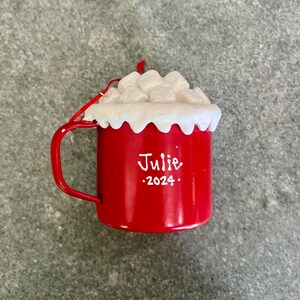 May include: A red enamel mug ornament with a handle, filled with white marshmallows and a white rim. The mug has the name "Julie" and the year "2024" in white script. A red string is attached for hanging. A festive holiday decoration.