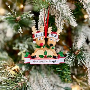 May include: A Christmas ornament featuring two reindeer with the names "Billy" and "Madi" on their hats. The reindeer are brown with red noses and green collars, and the ornament has a banner that reads "Happy Holidays."