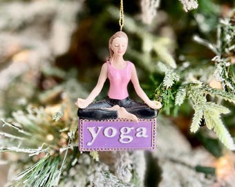 May include: A Christmas tree ornament featuring a woman in a yoga pose. The woman wears a pink top and black pants, seated in a lotus position. A purple sign below reads "yoga". The ornament hangs from a gold string.