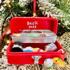 May include: A red Christmas ornament shaped like a tackle box, with the name "Beck" and the year "2023" on the lid. The tackle box contains miniature fishing lures and accessories. The ornament is hanging on a Christmas tree.
