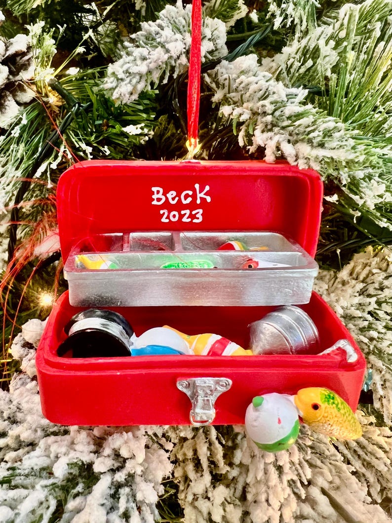 May include: A red Christmas ornament shaped like a tackle box, with the name "Beck" and the year "2023" on the lid. The tackle box contains miniature fishing lures and accessories. The ornament is hanging on a Christmas tree.
