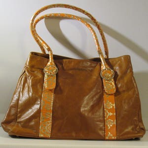 May include: A brown leather tote bag with orange and gold patterned straps. The bag has two top handles and a zipper closure.