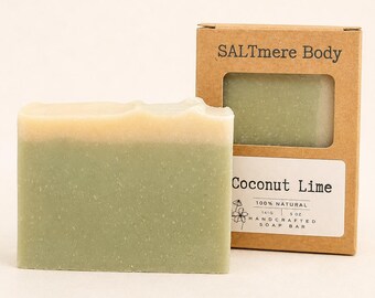 May include: A rectangular bar of soap with a light green base and a cream-colored top layer, positioned next to a brown cardboard box labeled SALTmere Body Coconut Lime, 100% natural, handcrafted soap bar, 141g, 5 oz, with a floral graphic.
