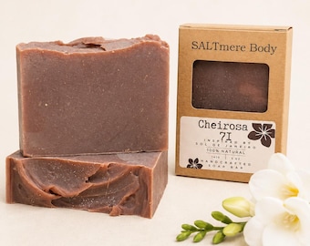 May include: Two rectangular bars of brown soap stacked next to a brown cardboard box labeled SALTmere Body Cheirosa 71. The label indicates 100% natural, 5 oz, and 141g. A small white flower with green buds sits in the foreground.