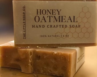 May include: Two bars of honey oatmeal soap are stacked on a wooden surface. The top bar has a paper label that reads "HONEY OATMEAL HAND CRAFTED SOAP 100% NATURAL 5 OZ." The soap is light brown with a textured top.