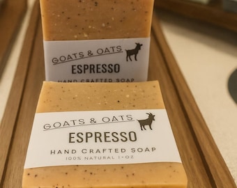 May include: Close-up of several rectangular bars of espresso-scented soap. Each bar is light brown with dark specks and a white label that reads "Goats & Oats Espresso Hand Crafted Soap 100% Natural 1+ oz."