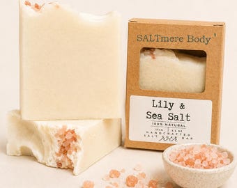 May include: Two off-white rectangular soap bars with visible pink salt crystals, one inside a brown cardboard box labeled SALTmere Body Lily & Sea Salt 155g 5.5 oz Handcrafted Salt Soap Bar, alongside a small bowl of pink salt crystals.