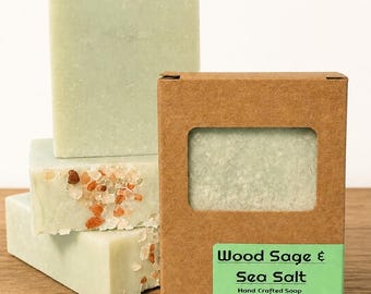 May include: Handmade green soap bars with sea salt crystals, stacked on a wooden surface. A brown cardboard box with a green label that reads "Wood Sage & Sea Salt" is next to the soap. The soap is 4.75 oz.