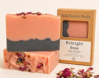 May include: Two rectangular bars of soap, one pink with a dark gray base and one pink with dried rose petals, next to a brown cardboard box labeled SALTmere Body Midnight Rose 100% Natural 141g 5 oz Handcrafted Vegan Soap Bar with rose buds.