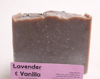 May include: A rectangular bar of handmade soap with a lavender and vanilla scent. The soap is a muted purple color with flecks of lavender and a pink label that reads "Lavender & Vanilla". The label also lists ingredients and the soap company.