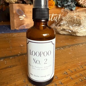 K&ouml;nnte beinhalten: Eine braune Glasspr&uuml;hflasche mit schwarzem Verschluss und wei&szlig;em Etikett. Auf dem Etikett steht "LOOPOO No. 2 Eau de Toilette. Literally 2oz., all natural" und "the little soap co. NEW ENGLAND". Die Flasche steht auf einer Holzoberfl&auml;che.
