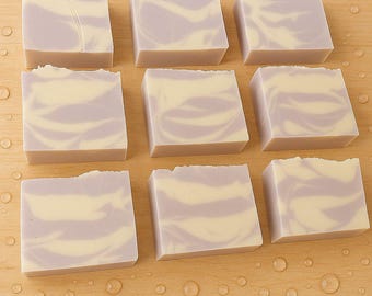 May include: Nine square bars of handmade soap with a lavender and cream swirl design. The soaps are arranged on a light wood surface with water droplets. The soap bars have a rustic, textured edge.