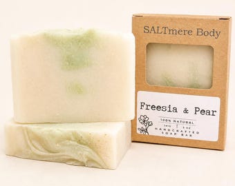 May include: Two rectangular bars of off-white soap with pale green swirls, accompanied by a brown cardboard box featuring a cutout window. The label reads SALTmere Body, Freesia & Pear, 100% NATURAL, 141G | 5 oz, HANDCRAFTED SOAP BAR.