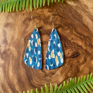 May include: A pair of blue and gold cork earrings with a geometric teardrop shape. The earrings have a white and gold pattern.