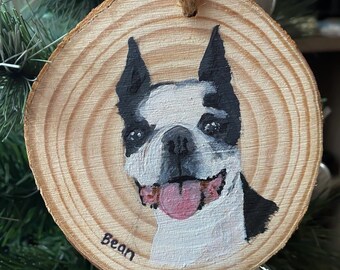 Puede incluir: Un adorno de madera pintado a mano con un perro Boston Terrier de pelo blanco y negro. El perro sonríe con la lengua fuera. El adorno es redondo y tiene un patrón de veta de madera. El nombre del perro, "Bean", está escrito en la parte inferior del adorno.