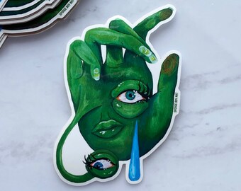 Puede incluir: Una mano verde con un ojo que llora y un segundo ojo en la parte inferior de la mano. La mano sostiene una lágrima azul. La pegatina tiene el texto "SPOOKY BOY ART".