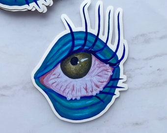 Puede incluir: Una pegatina de un ojo grande y azul con pestañas largas y oscuras. El ojo está dibujado en un estilo de dibujos animados y tiene una esclerótica blanca, un iris marrón y una pupila negra.