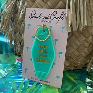 K&ouml;nnte beinhalten: Ein gr&uuml;ner Schl&uuml;sselanh&auml;nger mit einem goldenen Ring und dem Text "Meet me in Palm Springs" in goldenen Buchstaben. Der Schl&uuml;sselanh&auml;nger befindet sich auf einer rosa Karte mit dem Text "Scout and Craft Keychain" und dem Instagram-Handle "@scoutandcraftla".