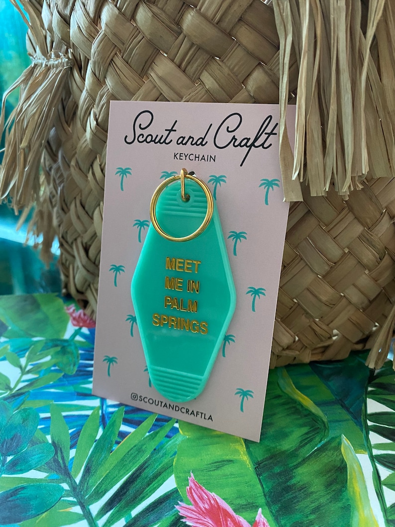 K&ouml;nnte beinhalten: Ein gr&uuml;ner Schl&uuml;sselanh&auml;nger mit einem goldenen Ring und dem Text "Meet me in Palm Springs" in goldenen Buchstaben. Der Schl&uuml;sselanh&auml;nger befindet sich auf einer rosa Karte mit dem Text "Scout and Craft Keychain" und dem Instagram-Handle "@scoutandcraftla".