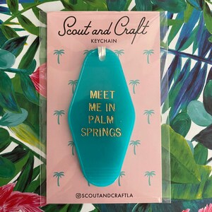 K&ouml;nnte beinhalten: T&uuml;rschild-Schl&uuml;sselanh&auml;nger in T&uuml;rkisblau mit goldener Aufschrift "Meet Me In Palm Springs". Der Schl&uuml;sselanh&auml;nger befindet sich auf einem rosa Hintergrund mit Palmenillustrationen.