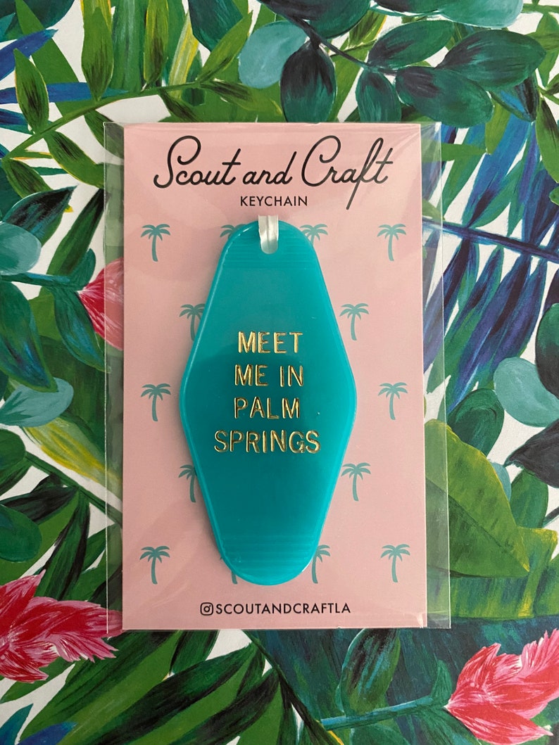 K&ouml;nnte beinhalten: T&uuml;rschild-Schl&uuml;sselanh&auml;nger in T&uuml;rkisblau mit goldener Aufschrift "Meet Me In Palm Springs". Der Schl&uuml;sselanh&auml;nger befindet sich auf einem rosa Hintergrund mit Palmenillustrationen.