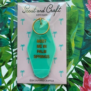 K&ouml;nnte beinhalten: Ein t&uuml;rkisblauer Motel-Schl&uuml;sselanh&auml;nger mit goldener Aufschrift "Meet Me In Palm Springs". Der Schl&uuml;sselanh&auml;nger ist auf einer rosa Karte verpackt mit den Worten "Scout and Craft Keychain" und dem Social-Media-Handle "@scoutandcraftla".