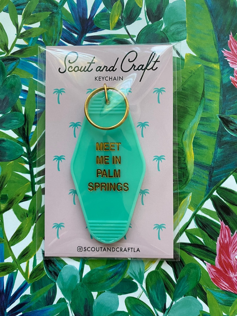 K&ouml;nnte beinhalten: Ein t&uuml;rkisblauer Motel-Schl&uuml;sselanh&auml;nger mit goldener Aufschrift "Meet Me In Palm Springs". Der Schl&uuml;sselanh&auml;nger ist auf einer rosa Karte verpackt mit den Worten "Scout and Craft Keychain" und dem Social-Media-Handle "@scoutandcraftla".