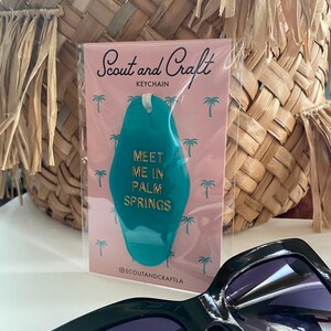 K&ouml;nnte beinhalten: T&uuml;rkiser Motel-Schl&uuml;sselanh&auml;nger mit goldener Aufschrift "Meet me in Palm Springs". Der Schl&uuml;sselanh&auml;nger ist auf einer rosa Karte mit aufgedruckten Palmen verpackt. Die Karte tr&auml;gt au&szlig;erdem den Text "Scout and Craft Keychain" und "@scoutandcraftla".