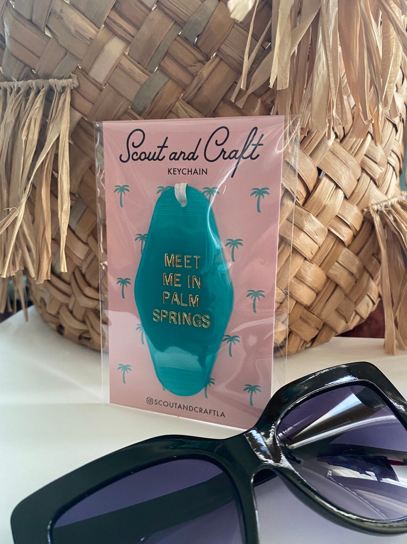 K&ouml;nnte beinhalten: T&uuml;rkiser Motel-Schl&uuml;sselanh&auml;nger mit goldener Aufschrift "Meet me in Palm Springs". Der Schl&uuml;sselanh&auml;nger ist auf einer rosa Karte mit aufgedruckten Palmen verpackt. Die Karte tr&auml;gt au&szlig;erdem den Text "Scout and Craft Keychain" und "@scoutandcraftla".