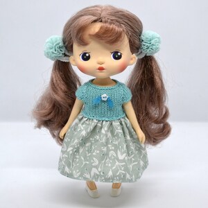 Może przedstawiać: Lalka z brązowymi włosami, niebieskimi oczami i sukienką w kolorze jasnoniebieskim i białym. Lalka ma na sobie jasnoniebieski sweter dzianinowy z niebieskim kwiatem z przodu. Lalka ma jasnoniebieską kokardkę we włosach.