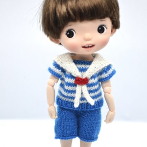 May include: A small doll with brown hair wearing a blue and white striped sailor shirt with a white collar and a red heart detail. The doll is wearing blue shorts and purple shoes.