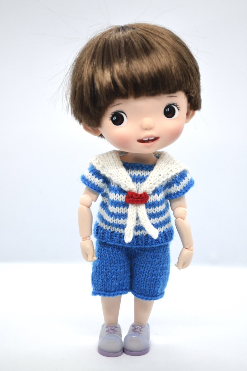 May include: A small doll with brown hair wearing a blue and white striped sailor shirt with a white collar and a red heart detail. The doll is wearing blue shorts and purple shoes.