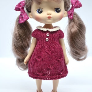 May include: A doll with long brown pigtails, each tied with a pink polka dot bow. The doll is dressed in a knitted burgundy dress with a white collar and two white buttons. Burgundy shoes complete the outfit.