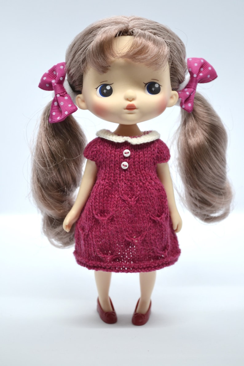 May include: A doll with long brown pigtails, each tied with a pink polka dot bow. The doll is dressed in a knitted burgundy dress with a white collar and two white buttons. Burgundy shoes complete the outfit.