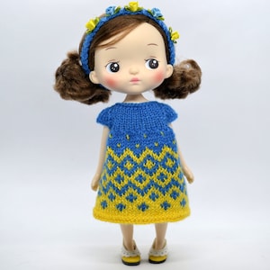 May include: A doll with brown hair styled in two pigtails, wearing a blue headband decorated with small yellow and blue fabric flowers. The doll is dressed in a short-sleeved knitted dress featuring a blue and yellow geometric diamond pattern.