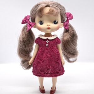 May include: A doll with long, light brown pigtails, each tied with a pink bow with white spots. The doll is dressed in a burgundy knitted dress with a white collar and red shoes. The dress has two white buttons.