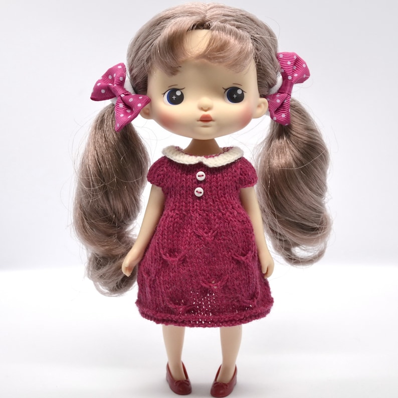 May include: A doll with long, light brown pigtails, each tied with a pink bow with white spots. The doll is dressed in a burgundy knitted dress with a white collar and red shoes. The dress has two white buttons.