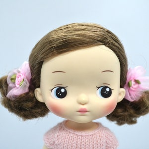 May include: A doll with brown hair, blue eyes, and rosy cheeks. She is wearing a pink knitted sweater and has pink bows in her hair.