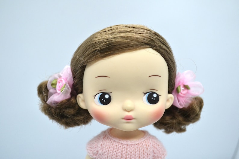 May include: A doll with brown hair, blue eyes, and rosy cheeks. She is wearing a pink knitted sweater and has pink bows in her hair.