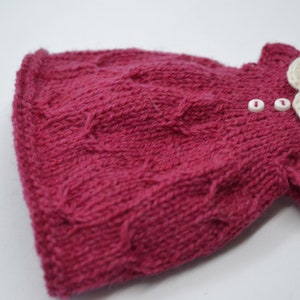 May include: A knitted magenta dress with a cable knit design. The dress has a white crocheted collar and two small white buttons. The garment is handmade, likely for a baby or small child.