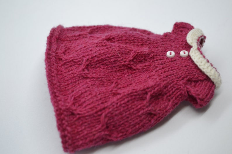 May include: A knitted magenta dress with a cable knit design. The dress has a white crocheted collar and two small white buttons. The garment is handmade, likely for a baby or small child.