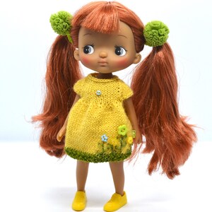 May include: A doll with long, red hair wearing a yellow knitted dress with green trim and floral accents. The doll has brown eyes and brown skin.